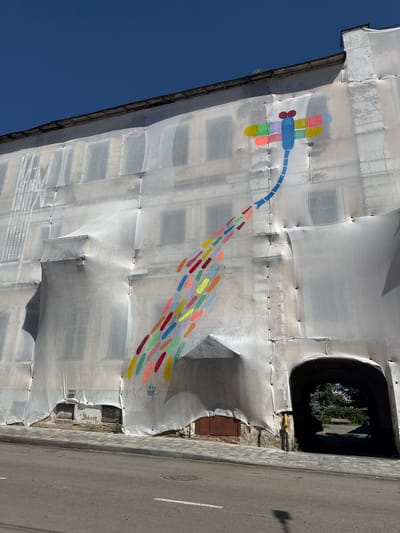 A street art mural of a colorful dragonfly with a trail of colored droplets extends across a building facade covered in white scaffolding sheeting in Yelets, Russia. The dragonfly, depicted in blue, red, yellow, green, and pink, is positioned at the top left of the building. Its body is blue, with red eyes and wings made of segmented colored rectangles. A trailing tail of numerous small, elongated droplets in various bright colors—including red, orange, yellow, green, light blue, and purple—extends downwards and to the right, creating a dynamic visual effect. The building itself appears to be under renovation or construction, as indicated by the extensive sheeting. The sheeting is draped over the windows and architectural details, creating folds and shadows. Below the main facade, there is an arched entryway, and the street in front is paved with asphalt and bordered by a sidewalk made of stone pavers. The sky is clear and blue.