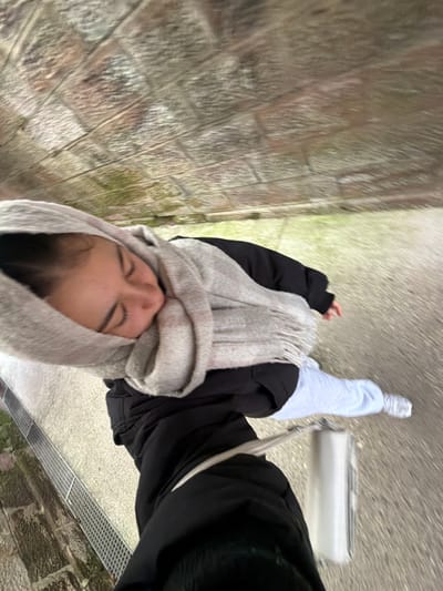 The image captures a first-person, slightly downward-angled perspective of a female subject walking outdoors in Obermodern-Zutzendorf, France. The subject's head and upper torso are visible, with her eyes closed or looking downwards, wearing a light grey scarf wrapped around her head and neck, over a black jacket. Below the jacket, light blue sweatpants and white sneakers are visible. A white rectangular bag with a white strap hangs near her lower right side. To the left, a metal drainage grate runs parallel to the walking surface. The ground consists of a light-colored paved path with patches of green growth. Above the subject, a curved, vaulted ceiling or wall constructed from irregular, reddish-brown and grey stones is visible. The scene suggests casual movement in an outdoor, possibly underpass or archway, setting.