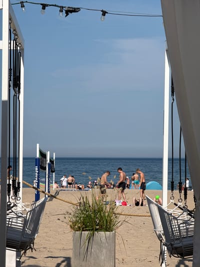 The image shows a sunny day at a beach in Sopot, Poland, likely near the pier, as indicated by the presence of beach volleyball nets and a relaxed, vacation atmosphere. In the foreground, white metal chairs hang from a white frame, adorned with black ropes. A concrete planter with tall, feathery grass sits to the right of the chairs, casting a shadow on the sandy ground.
The middle ground features a beach with several groups of people. Some are standing and interacting, while others are sitting or lying down. Two volleyball nets with green mesh and blue poles are visible, suggesting recreational activities. A blue tent is pitched on the sand. The ocean, calm and blue, forms the background, meeting a clear, pale blue sky. String lights are strung across the top of the frame, indicating an outdoor seating or relaxation area, possibly for a nearby establishment. The overall scene is one of leisure and summer enjoyment.