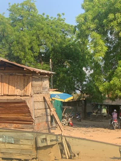 This image depicts an outdoor scene in Abobo, Nigeria, characterized by rudimentary structures and natural vegetation under clear daylight conditions.
In the left foreground, a two-story structure constructed from raw, horizontally laid wooden planks with a corrugated metal roof is visible. The lower portion of this structure appears partially obscured by a reflective surface, possibly a vehicle window. A rough-hewn wooden beam leans diagonally against the side of this building.
The midground features an unpaved, sandy-dirt surface. A blue and yellow umbrella, likely marking a vendor's stall, stands above a small green corrugated metal structure. Beneath this umbrella, a red and silver motorcycle is parked. Further into the midground, another motorcycle is parked. Towards the right, a person wearing a purple long-sleeved shirt and a white cap is seated on a motorcycle, facing away from the viewer and apparently in motion or about to move. Behind these vehicles and people, additional simple structures built with corrugated metal and wooden planks are visible, suggesting dwellings or small businesses.
The upper portion of the image is dominated by dense, green tree foliage against a light blue sky, indicating lush vegetation in the area. A utility pole with visible wires extends vertically near the center-left. The scene is brightly illuminated by natural sunlight.