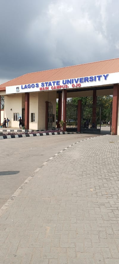 The image depicts the main entrance to Lagos State University, Main Campus, located in Ojo, Lagos State, Nigeria. The scene is observed under an overcast sky filled with grey clouds.
In the foreground, a light grey interlocking paver surface extends from the lower right towards the center, meeting an asphalt road on the left. A curb, striped black and white and lined with small reddish-purple plants, separates these two surfaces and curves towards the entrance structure.
The central feature is a large entrance gate structure topped with a terracotta-colored, ridged roof. Below the roofline, a prominent blue sign reads "LAGOS STATE UNIVERSITY MAIN CAMPUS, OJO," accompanied by a university emblem on the left side of the text. The structure is supported by multiple dark brown pillars.
To the left of the main archway, a light-beige building, possibly a guardhouse or administrative office, features several small rectangular windows. Four individuals are present near this building: one person, dressed in dark clothing, stands facing right with an object on their head; three others are clustered further left, two standing and one appearing to lean against a surface.
Beyond the main archway, dark, decorative metal gates are partially visible. Through the gates, green foliage from trees and shrubs, along with distant paved areas, indicate the interior of the campus. On the right side of the structure, another brown pillar and a potted plant are visible. No explicit actions or interactions beyond the individuals' stationary presence are discernible.