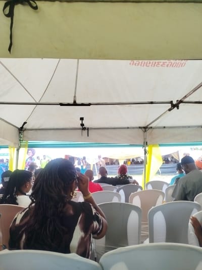 The image shows a gathering of people in what appears to be an event in Ukwulu, Nigeria. The scene is viewed from under a white tent, with rows of white plastic chairs occupied by attendees. Many of the people in attendance are facing a stage or presentation area, located beyond the tent's opening. Several individuals are visible on the stage, and some people are standing in front of the stage as well. The attendees appear to be observing a presentation.