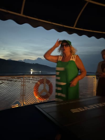 The image shows a woman wearing a green dress and a green hat on a boat at dusk. She is standing on the deck of the boat, with her right hand raised to her hat. The woman is wearing sunglasses and has blonde curly hair. Behind her, another person is visible. The boat is on a body of water, with a shoreline and hills in the background. The sky is cloudy and dark. There is a life preserver hanging on a fence in the foreground. The text on the table in the foreground appears to be in Russian and reads "БАЛТИКА". The city of Гольяны is located on the banks of the Volga River, and it is possible this image was taken on a boat on the Volga River near Гольяны, Russia.