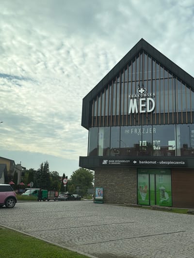 The image shows a modern building in Limanowa, Poland, with the signage "Krakowska Med" visible on its upper facade. Below this, the text "FRYZJER" (Polish for hairdresser) is displayed across the glass windows. The ground floor features signage for "BANK SPÓŁDZIELCZY W LIMANOWEJ" (Cooperative Bank in Limanowa) and "bankomat - ubezpieczenia" (ATM - insurance). In front of the building, a paved area transitions to a grassy strip. Several vehicles are present, including a pink Mini Cooper, a green van pulling a trailer, and other cars. People are visible near the vehicles. The sky is cloudy. The building's architecture is contemporary, with a dark facade and vertical wooden accents. To the right of the main building, a storefront displays images of hot dogs and coffee, with the text "Głodny?" (Hungry?) and "Gorąca kawa?" (Hot coffee?) visible.