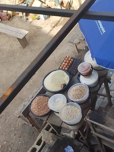 The image depicts an outdoor market scene in Abeere, Nigeria. In the foreground, various food items are displayed on a rustic wooden table. There are containers filled with different types of grains, including what appears to be rice and smaller seeds. A tray of brown eggs is also visible, arranged neatly. The setting is informal, with the ground being dirt and scattered debris. In the background, there are cardboard boxes and remnants of what might be a stall or shop. A blue umbrella, possibly advertising "Moniepoint Microfinance Bank", is positioned to the right, suggesting a commercial area. A wooden bench is visible further back. The lighting suggests it is daytime, and the overall impression is that of a busy market environment with goods being prepared for sale. There is no direct indication of weather conditions, but the open-air setting implies fair weather. The focus is on the agricultural products and the basic setup of a local market stall. The text "Moniepoint Microfinance Bank" is clearly legible on the umbrella.
