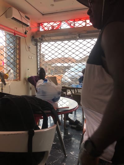The image depicts an indoor scene in Ede, Nigeria, characterized by an apparent waiting or commercial area. In the foreground-right, a person is partially visible in profile, wearing a dark cap, a black and white sleeveless top, and a watch on their right wrist. In the midground-left, an adult male with dark skin and a shaved head, dressed in a purple long-sleeved shirt, sits in a chair, leaning back and looking towards the camera.

Several objects are present: A round, glass-topped table with a red base is in the midground, holding a large plastic water bottle contained within a clear plastic bag labeled "EMMA." A black backpack rests on a white chair in the immediate foreground. Another round table with smaller bottled water is visible in the background. The floor is dark-colored tile with a marbled pattern. On the left wall, a white wall-mounted air conditioning unit is installed, and below it, a small fan with yellow blades is visible near a window. Both the window and the main opening to the exterior are secured with metal grilles. Red fluorescent lighting or decorative elements are positioned above the main opening. Outside the grilles, an indistinct urban environment with faint vehicle shapes is visible, suggesting a street or public space during daylight hours. A pair of black slide sandals rest on the floor beneath the midground table.