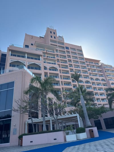 A large, multi-story building, likely a hotel or residential complex, dominates the scene under a clear blue sky. The building is a light pinkish-beige or sandy color, featuring a stepped architectural design that recedes at various levels. Its facade is characterized by numerous rectangular windows and recessed balconies. Some balconies are adorned with intricate white geometric latticework and arched screens, while others have modern glass railings.
Several tall palm trees are strategically placed around the building, with their trunks and lush green fronds visible, enhancing the tropical resort atmosphere. The lighting suggests bright daylight, possibly mid-morning or early afternoon, with a strong sun glare visible towards the upper right of the image. The sky is entirely clear and cloudless.
At ground level, there are paved areas, including a prominent strip of vibrant blue, which could be part of a walkway or pool edging, juxtaposed against textured grey tiles. Smaller architectural elements, possibly outdoor lounges or amenity structures, with white walls and a visible wooden bench, are also present. Sparse greenery, including small bushes and additional trees, contributes to the landscaping.
On a lower section of the building, a security camera is mounted, and a rectangular plaque or sign is affixed to the wall, though its text is not legible. The scene is devoid of people or discernible activity, conveying a tranquil and pristine environment. The overall aesthetic, with its distinctive architecture and palm trees, strongly aligns with the context of a luxurious development in a warm climate, such as Al Fiqait, United Arab Emirates.