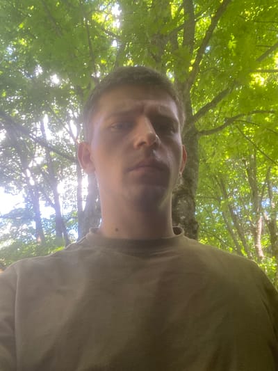A man is standing outside under a canopy of green leaves. The man is wearing a brown t-shirt. The leaves are lit by sunlight, giving the scene a bright, natural appearance. The background is a forest with a lot of trees. The man is looking at the camera. The location is unknown.