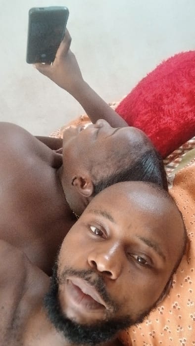 Two men lie side-by-side. One man, with a full beard, looks into the camera. The other man has a shaved head and is angled away from the camera. The man holds a cell phone in his hand. They lie on top of patterned bedding.