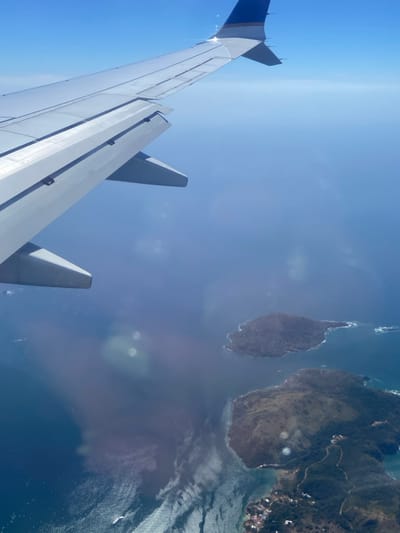 An aerial photograph captures a segment of a silver aircraft wing in the upper left, featuring a blue and white livery accent on its wingtip and visible flap components. The scene below depicts a clear blue ocean body interspersed with two distinct landmasses, characteristic of the coastal area around Santa Cruz de Chinina, Panama. The larger island in the lower right displays varied terrain, including dense green vegetation, cleared patches, and approximately five discernible residential structures connected by a visible dirt road along its western coastline. A smaller, densely vegetated island appears further out in the water towards the image center. The water exhibits varying shades of blue, indicating depth changes, with white wave patterns visible near the shorelines and a small white boat situated in the lower-left quadrant. The sky is uniformly clear and bright blue, filling the upper-right portion of the frame. Sunlight reflections are present as circular lens flares on the viewing surface. No individuals are visible.
