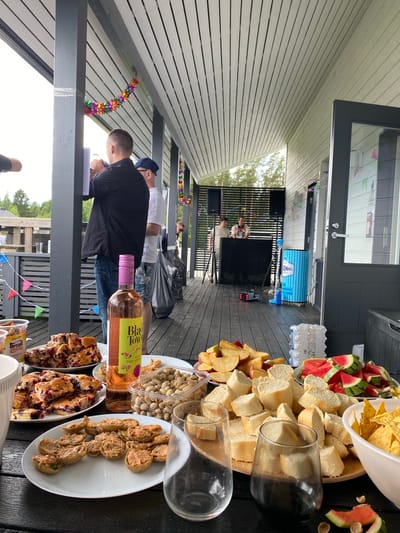 This image captures an outdoor gathering, likely a party or casual get-together, set on a wooden deck with a covered roof. Several people are present, some standing and interacting, while others are engaged in activities like setting up equipment or preparing food. In the background, a person appears to be setting up audio equipment, with speakers and a large black speaker visible. There are also signs of a celebration with colorful garland strung across the area and small triangular flags.
A prominent feature of the scene is the spread of food and drinks laid out on tables. This includes platters of bread, chips, pistachios, and what appear to be some sort of pastry or dessert. A bottle of rosé wine with a label reading "Black Tower" and mentioning "Pink Grape" is clearly visible. Other food items include sliced watermelon, and what looks like baked goods with berries. A large roll of paper towels or similar item is also present, along with a blue cylindrical object that might be a cooler or a piece of equipment, with text on it that includes "GINAL" and "a 1952".
The lighting suggests it is daytime, likely a sunny afternoon, given the shadows and bright illumination. The setting appears to be a residential area, possibly a backyard or patio of a house, indicated by the decking and the natural greenery visible beyond the structure. The overall mood seems relaxed and festive.