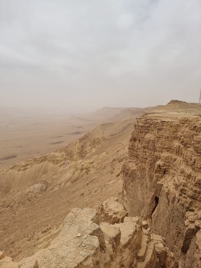 The image shows a rocky desert landscape in Mitzpe Ramon, Israel, possibly a cliff or canyon. The sky is overcast with a thick, hazy cloud cover. The rock formations are primarily light brown and beige, with visible layers and textures indicating sedimentary rock. The landscape is arid and barren.
