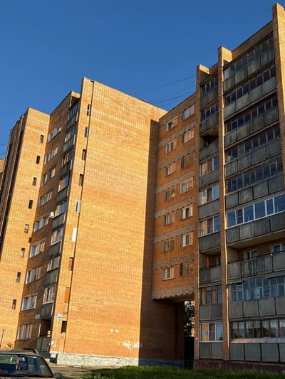 This is an exterior, low-angle, vertical-format, medium shot of an old, multi-story brick apartment building under a clear blue sky. The building is constructed of orange-brown bricks and features numerous balconies with gray, weathered railings. Many of the windows are visible, some reflecting the sunlight. A passage or archway is present between sections of the building, revealing a glimpse of trees and foliage beyond. The building appears to be located in a residential area, with a patch of grass and a portion of a car visible in the foreground. There are no people in the frame. The building's architecture suggests it is likely from the Soviet era, typical of many buildings found in cities like Kirovo-Chepetsk, Russia.