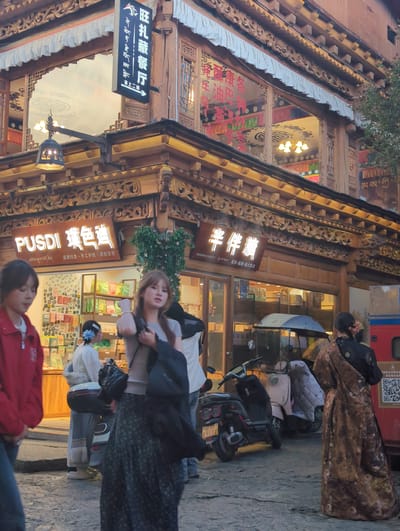The image captures a street scene in the Old Town of Shangri-La, China. The architecture features traditional Tibetan wooden carvings and overhangs, with several shops displaying their signs. One sign, in large white lettering, reads "PUSDI" followed by Tibetan script and smaller Chinese characters indicating "Tibetan Cuisine." Another sign, also in Tibetan and Chinese, reads "BAN BAN SWEETY" and advertises "Tibetan Tea, Hand-made pastries, Creative gifts."

In the foreground, a young woman in a red jacket and jeans walks past a shop. To her right, another young woman in a grey top and a long patterned skirt is walking and holding a black bag. Behind them, a person in traditional white and grey Tibetan attire with fur trim stands near a motorbike. Further back, a person wearing a richly patterned golden and black traditional robe walks away from the camera. Several motorbikes and a small pink scooter are parked on the cobblestone street. The lighting suggests it is late afternoon or early evening, with warm light illuminating the scene and casting soft shadows.
