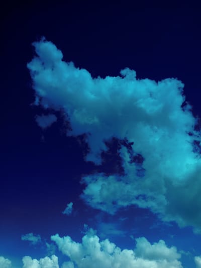 The image displays a dramatic, low-angle view of a dark blue sky filled with wispy, cyan-tinted clouds. The clouds are concentrated in the upper and central portions of the frame, with a larger, more defined cloud formation dominating the upper right quadrant. These clouds appear to be illuminated from an unseen source, creating a striking contrast against the deep blue of the sky. A few scattered, lighter clouds are visible towards the bottom of the image, appearing more white and less intensely colored. The overall scene evokes a sense of atmosphere and depth, with the dark sky suggesting twilight or a stormy condition. There are no people, objects, or discernible urban elements present to indicate a specific location within Voznesenovka, Russia.