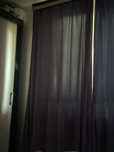 The image depicts an interior scene with a tall, narrow window partially obscured by dark purple, sheer curtains. The curtains are pleated and hang from a rod at the top, casting subtle shadows. To the left of the curtains, a portion of a white sliding closet door is visible, with a sleek, horizontal chrome handle. Above the closet door, on a shelf, are a few objects including what appears to be a white plastic bag and some cardboard packaging. The wall behind the closet door and to the right of it has a textured wallpaper with a subtle pattern. The lighting suggests an indoor environment, possibly a bedroom or living area. This scene could be located in Mirnyy, Russia, a city known for its diamond mining industry and consequently, residential buildings. The overall impression is one of a quiet, domestic space.