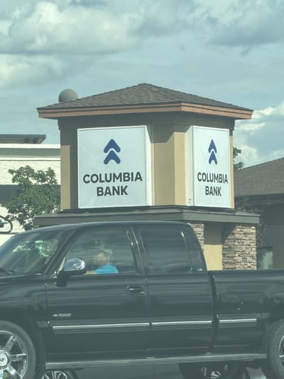 A black pickup truck is parked in front of a Columbia Bank building. The truck has tinted windows, chrome wheels, and "1500" emblem on the side. A person wearing a blue shirt and a baseball cap is visible in the driver's seat. The bank building is light tan with a brown roof and has two signs with the Columbia Bank logo and name. The background shows a cloudy sky and trees. The scene is located in Pasco, United States of America.