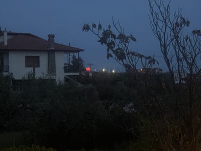 The image depicts a nighttime scene in Γερακινή, Greece. In the foreground, a tree with sparse leaves obscures the view. Behind the tree, a white two-story building with a red-tiled roof is visible. Further in the background, a red sign is illuminated, possibly indicating a business. Streetlights and distant buildings can be seen in the background, suggesting a residential or commercial area. The overall scene is dark and the details are obscured by the darkness.