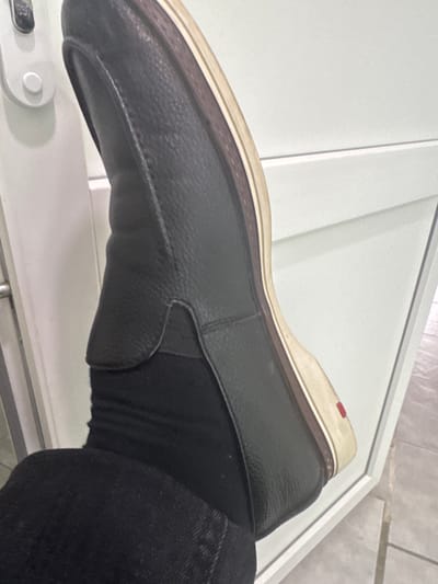A single black leather loafer is on a foot wearing black socks and dark denim jeans. The foot and shoe are positioned near a white surface, likely a door or wall with a white frame. The setting appears to be indoors, potentially within a residential or commercial building. The background includes neutral-colored tiled surfaces. The location in the city of Bakhchysarai, Ukraine is not possible to confirm from the image.