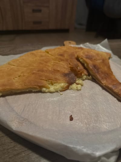 A golden-brown pie, possibly khachapuri, rests on white parchment paper. The crust is uneven and slightly charred in places, revealing a creamy, pale filling along the exposed edge. A slice has been removed from the top. A small crumb rests on the paper to the right. The scene is set on a wood surface, and a cabinet is partially visible in the background.