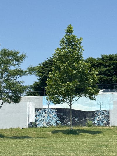 The media file shows a mural painted on a concrete wall, with a tree in the foreground and a grassy area to the right. The mural features stylized trees with blue and white ornamental patterns, set against a dark background. The scene appears to be outdoors, with a clear blue sky visible above the trees. The lighting suggests it is daytime, likely sunny given the shadows cast by the trees and the bright grass. The location is identified as Buffalo, United States of America. No people are visible in the frame, and there is no discernible text within the image. The overall impression is a peaceful, perhaps urban, outdoor setting enhanced by the artistic mural. The composition is vertical, with the wall and mural occupying the right half of the frame and the sky and foliage dominating the left.