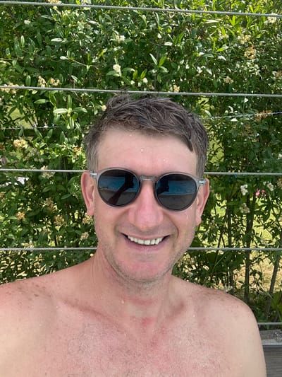 A close-up, low-angle selfie shows a person from the chest up, with their head tilted back and facing the camera. They are wearing dark, round sunglasses and have wet, dark hair. The person is smiling broadly, revealing their teeth. Their skin is pink and appears to have water droplets on it, suggesting they may have been swimming or are in a humid environment.

The background is dominated by lush green foliage, with some small white flowers visible. Thin, metallic wires run vertically through the frame, partially obscuring the greenery. The lighting suggests it is daytime, possibly sunny. The overall mood is relaxed and happy.

The image was taken in Saint-Imier, Switzerland. There is no visible text in the image.