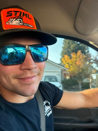 A male subject is depicted from the chest up, seated in the driver's position within a vehicle on a residential street in Rancho Murieta, United States. He wears a black baseball cap with an orange patch featuring the word "STIHL" and a chainsaw graphic, dark-framed blue mirrored sunglasses, and a dark t-shirt with a visible 'Memorial 2021' graphic on the left chest. A light brown mustache is present above his upper lip. The subject displays a slight smile, with his head tilted slightly to the right. He is secured by a grey seatbelt diagonally across his chest and left shoulder. His left arm is visible, resting on the dark interior door panel of the vehicle. A beige headliner is visible in the upper portion of the vehicle's interior. The subject's left sunglass lens reflects another individual, likely the photographer, holding a smartphone while seated in the passenger seat. Through the vehicle's window, a clear daylight scene of a residential area is visible. In the background are single-family houses, one with a prominent white garage door, and trees displaying both green and autumnal yellow-orange foliage. A white vehicle is partially visible parked or in motion in the background.