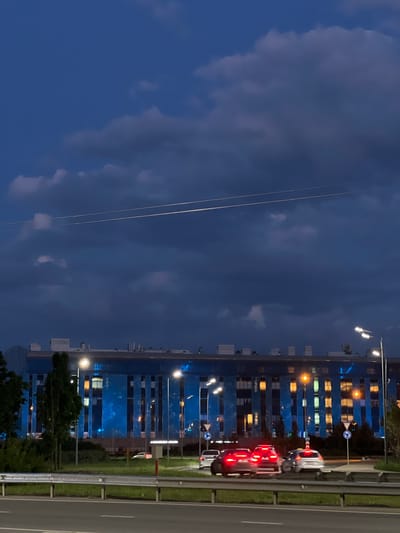 An exterior shot of a blue-paneled building in Dachnoe, Russia, is captured at dusk. The building features numerous windows, some of which are illuminated with warm-toned light, contrasting with the cool blue exterior. Several streetlights are visible, casting a glow on the scene. A highway in the foreground shows a few cars in motion, their headlights and taillights creating streaks of light due to the long exposure. The sky above is a deep blue, with a few scattered clouds. Trees and greenery line the area between the highway and the building.