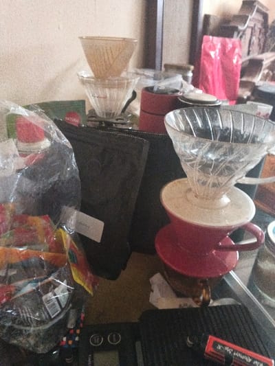 The image depicts a static indoor scene on a cluttered surface, likely a table or counter, in Dogsenang Gorphoog, Bhutan. The primary subjects are various items associated with manual coffee brewing.
In the center, two conical pour-over coffee drippers are stacked; the upper one is an amber-toned plastic or glass, resting on a clear ribbed glass dripper, which is positioned on a black stand or scale. To the right, a clear ribbed glass dripper sits on a white ceramic collar, which then rests on a red ceramic mug or brewer with a handle.
To the far left in the foreground, a transparent plastic bag contains multiple individually packaged, colorful snack items. A small, white rectangular label is affixed to this bag. Behind this, a flat black pouch or bag stands upright.
In the bottom center, a black digital scale, partially legible as "TIMEMORE" or similar, is present on a clear glass surface. To its immediate right, a small red rectangular box with white lettering is also on the glass surface.
In the mid-ground, behind the central stacked drippers, a stack of red cylindrical objects, possibly a manual coffee grinder or storage containers, is visible. Further in the background, a bright pink or red flexible bag, likely containing coffee beans, stands upright. The background consists of a light-colored wall and indistinct darker elements, possibly furniture or shelves. No individuals are present in the scene, and no explicit actions are occurring. The environment suggests an active coffee preparation area.