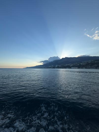 The image shows a view of the sea and a mountain range in Гаспринский поселковый совет, Ukraine. The sea is on the right, with dark blue water and some white foam visible. The mountain range is on the left, with several buildings visible on the slopes. The sky is blue, with some clouds, and the sun is shining behind the mountains, creating a ray effect.