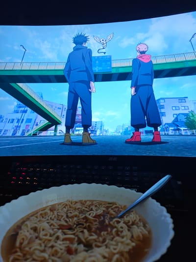 The image depicts a television screen displaying an anime scene featuring two male characters standing on a street, facing away from the camera. One character has spiky black hair and wears a dark blue jacket and pants. The other character has blonde hair and wears a red hoodie under a dark blue jacket and pants. Both characters are wearing matching brown shoes. In the background, there is a green elevated walkway or bridge with buildings visible beyond it. A small, winged creature with a skull-like head hovers in the sky between the two characters. Below the screen, a keyboard with illuminated keys is visible, and in the foreground, a bowl of ramen noodles with a spoon is placed on the keyboard. The scene appears to be set in a city. There is no indication of the city being Оқжетпес, Kazakhstan.