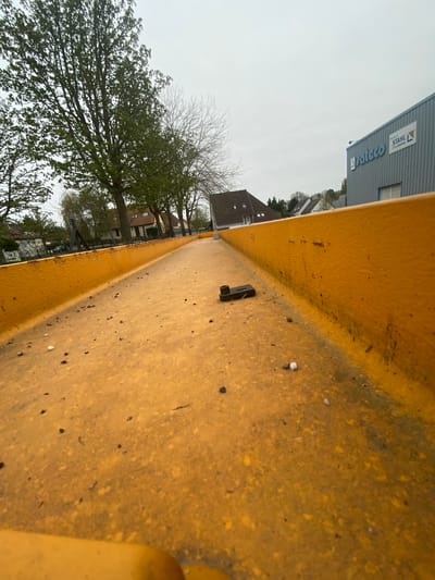 The image shows a close-up view of the interior of a yellow, potentially metal, container or structure. Debris, including small dark particles and a single white pellet, is scattered on the surface. A small, black, rectangular object with a raised feature is visible.
In the background, out of focus, is a street scene in Truchtersheim, France. Buildings, including houses with pitched roofs and a commercial building with "Stahlco" signage, are visible. Trees, some with sparse foliage, are present. The sky is overcast and grey.