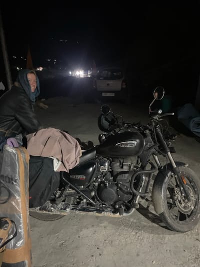 A person is lying down, partially covered by a blue scarf, next to a parked Royal Enfield Meteor 350 motorcycle. The motorcycle is black with chrome accents and has "Royal Enfield" and "Meteor 350" visible on its fuel tank and side panel respectively. The scene is dimly lit, suggesting it is nighttime or indoors in a dark area. The ground appears to be concrete or a similar rough surface. In the background, there is a body of water with lights reflecting on it, and a structure that could be a building or a boat with lights. The person's expression is relaxed and they appear to be resting. The overall setting seems to be outdoors in a somewhat rural or industrial area, possibly near a waterfront. The location context is Khangkor, India.