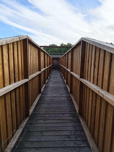 The image depicts a wooden walkway, possibly part of a boardwalk or an elevated path, leading towards a distant structure. The walkway is flanked by tall, vertical wooden planks that form walls on either side, creating a symmetrical perspective. The planks are a warm, natural wood color, and some appear weathered. The walkway itself is constructed from dark, horizontal wooden planks, showing signs of wear and tear. In the background, trees and a sliver of a building are visible, suggesting a natural or park-like setting. Above, the sky is a vibrant blue with wispy white clouds, indicating daytime. The scene appears to be outdoors, in a tranquil environment, possibly in the city of Slaka, Sweden, given the prompt's context. There are no people or animals visible in the image.