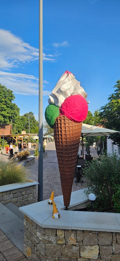 A large, three-scoop ice cream cone sculpture stands in a paved outdoor area in Balatongyörök, Hungary. The sculpture features a brown waffle cone with white whipped cream, a pink scoop, and a green scoop. Two small gnome figurines, wearing cone-shaped hats, are positioned on a stone ledge in the foreground. In the background, there are trees, buildings with awnings, outdoor seating areas, and several people walking. The sky is blue with scattered clouds.