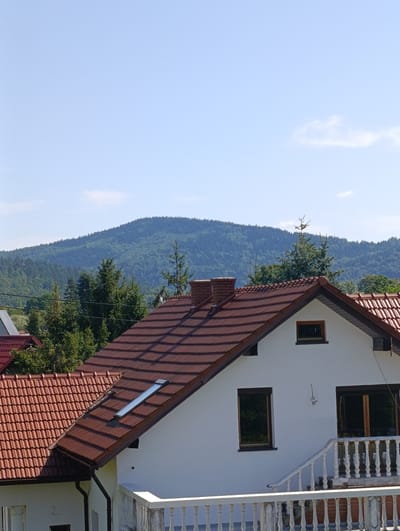 A bright, clear sky with a few wispy clouds forms the background of this outdoor scene. In the middle ground, a lush, green, forested mountain range stretches across the horizon. In the foreground, the tiled roof of a white, two-story house is visible. The roof is a deep reddish-brown color and features a chimney and a skylight. A white balcony with a railing overlooks the scene. The house appears to be situated in a residential area, possibly on the outskirts of Sucha Beskidzka, Poland, given the mountainous backdrop. The overall impression is one of a peaceful, rural setting on a sunny day.