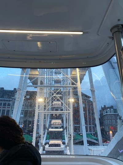 The image captures a view from inside a modern Ferris wheel gondola, looking outwards through a rain-splashed window. The perspective is tilted, offering a dynamic glimpse of the surroundings.
Inside the gondola, a clean, white interior wall is visible on the left, featuring a bright, vertical strip light. A metallic handrail or support bar runs along the top edge of the frame. In the bottom right corner, the back of a person's head and shoulder, with dark, curly hair, is visible, indicating they are also looking out.
The exterior view is dominated by the intricate white metal structure of the Ferris wheel itself. Numerous raindrops cling to the transparent window, causing slight distortions of the scene outside. Several other gondolas are visible, suspended from the Ferris wheel's frame, with some interior lights reflecting. The word "OVERVIEW" is clearly visible on at least two of these distant gondolas.
Beyond the Ferris wheel, parts of urban buildings with architectural details can be discerned, suggesting an urban setting, specifically Luik (Liège), Belgium, as per the context. The lighting suggests dusk or night, with artificial lights illuminating the Ferris wheel and reflecting off the wet surfaces. The raindrops on the window indicate either current precipitation or recent rain, contributing to a moody, atmospheric setting of an evening Ferris wheel ride.