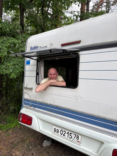 An adult male is prominently featured, leaning out of the open window of a white Hobby De Luxe camper van, which is stationary in a wooded outdoor setting in Fort-Krasnaya Gorka, Russia. The man, with a bald head and light stubble, wears a pale yellow short-sleeved t-shirt and smiles directly at the viewer, resting his forearms on the window's lower frame.

The camper van displays a textured white exterior with two blue horizontal stripes along its lower half and "Hobby De Luxe" branding on its upper rear. A dark red horizontal light bar is visible near the roofline, and a rectangular blue sticker is affixed to the side above the window. The vehicle exhibits signs of wear, including dirt accumulation, minor surface damage, and a weathered taillight on the lower left. A Russian license plate, "BB 0215 78 RUS," is mounted centrally on the rear bumper, with "78" indicating the Saint Petersburg region. A grey support block is positioned beneath the rear bumper.

The surrounding environment consists of dense green deciduous trees and foliage in the background, suggesting a natural, possibly forested area. The ground beneath the camper is composed of dirt and scattered organic debris, with sparse green ground cover. The lighting suggests daytime conditions, with diffused illumination.
