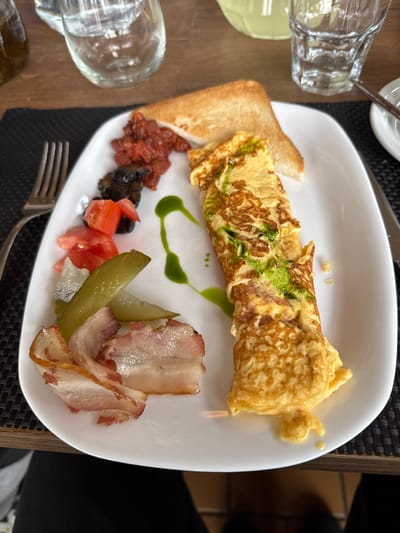 The image shows a breakfast plate with an omelet, toast, bacon, pickles, tomatoes, and olives. There is a green sauce drizzled on the omelet and on the plate. The breakfast is served on a white plate with a dark placemat. In the background, there are glasses of water and a blurred background that appears to be a table setting. The location is not specified in the image, and therefore it cannot be determined to be in Слабадскі сельскі Савет, Belarus.