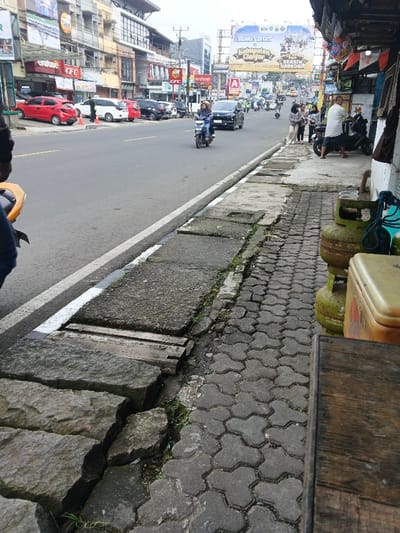 The image depicts a bustling street scene in Cianjur Regency, Indonesia, under an overcast sky. The viewpoint is from a low angle on the right sidewalk, looking down a two-lane asphalt road that recedes into the distance.
In the immediate foreground on the right, a dark, worn wooden table surface is visible. To its left, two stacked, weathered green LPG gas cylinders show signs of corrosion, accompanied by a yellow-cream insulated cooler box and a coiled blue rope. The sidewalk directly beneath these objects is constructed from interlocking hexagonal concrete pavers, which are worn, dirty, and have small patches of grass growing in the crevices. Further left, closer to the road, the sidewalk transitions to a section of large, uneven stone slabs, deeply cracked, with two wooden planks bridging a significant gap over what appears to be a drainage channel. The edge of the road is marked by a solid white line.
On the road, traffic is in motion. A person wearing a helmet rides a dark scooter in the middle lane, positioned behind a dark sedan. Further down the road, multiple vehicles, including cars and motorcycles, are visible, some moving and some parked.
The left side of the street features several commercial buildings, typically two to three stories high, with businesses such as "CFC" (California Fried Chicken) and "erofone" visible through their signage. Parked cars, including a red hatchback, a white sedan, and a black SUV, line the curb.
On the right side of the road in the midground, a man in a white t-shirt stands next to a parked black motorcycle on the sidewalk. A group of at least three women are also visible further down the right sidewalk.
In the background, a large billboard spans the street, displaying the Indonesian text "SELAMAT DATANG DI KABUPATEN CIANJUR" (Welcome to Cianjur Regency) and promoting a "BERSIN NARKOBA" (Drug-Free) campaign. Beyond the billboard, more buildings and distant, hazy hills are visible under the cloudy sky. Utility poles and wires crisscross the upper part of the scene.