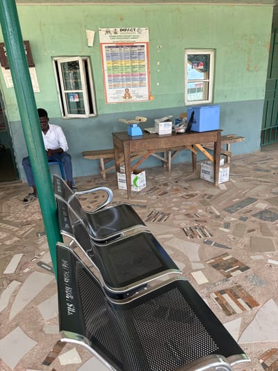 The image depicts the waiting area of a health facility in Lau, Nigeria. A man in a white shirt and blue pants sits on a blue chair, looking at his phone. In front of him are several rows of black metal chairs with perforated seats and chrome frames. A green pole is visible in the foreground. A wooden table with a blue scale, medical supplies in boxes, and a blue cooler box is set up against a light blue and green wall. The wall has two windows, one of which has a poster with medical information. The floor is covered with a mosaic of broken tiles. A green metal gate is visible in the background.