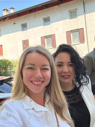 Two women are posing for a selfie in front of a building. The woman on the left has blonde hair and is wearing a white embroidered shirt. The woman on the right has dark curly hair and is wearing a black lace top and a white pinstripe blazer. The building in the background is light-colored with red-brown shutters on the windows. The scene appears to be in a city, possibly Udine, Italy, given the architectural style. The lighting suggests it is daytime, with sunlight casting shadows. There are also some trees visible to the left of the women.