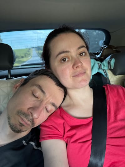 A man and a woman are in the back seat of a car. The man is asleep, with his head resting on the woman's shoulder. He has a goatee and is wearing a black t-shirt with white stripes on the sleeve. The woman is looking at the camera with her mouth slightly open. She is wearing a pink t-shirt and a black seatbelt. In the background, a road can be seen stretching into the distance, with trees and fields on either side. The image appears to be taken in daylight.