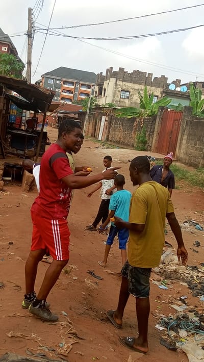 The image depicts an outdoor street scene in Awada, Nigeria, on a reddish-brown dirt road under an overcast sky.

In the foreground, a dark-complexioned adult male, wearing a red short-sleeved athletic shirt with black accents and red athletic shorts with white stripes, is actively speaking with his mouth open. His right arm is extended, pointing towards the right, and he holds a small, yellowish object in his left hand. To his right, a second dark-complexioned adult male, dressed in a mustard yellow short-sleeved t-shirt and dark patterned shorts, walks towards the left, holding a similar yellowish object in his right hand.

Behind these two primary figures, three additional individuals are visible. A young male in a light-colored patterned shirt points with his right hand towards the right. Nearby, a younger male in a light blue t-shirt and dark blue shorts stands. Further in the background, another adult male, wearing a dark striped long-sleeved shirt and a pink and white knitted cap, observes the scene.

The unpaved ground is uneven and extensively littered with various plastic debris, wrappers, and general refuse. To the left, a rudimentary stall or shop structure is visible, constructed with a wooden frame and corrugated metal roof, with stacked blue, red, and yellow plastic crates. An open ground access point, possibly a manhole or drainage, is situated near this stall. To the right, a low concrete wall, topped with banana plants and featuring a rusted metal gate, parallels the road. A significant pile of trash and discarded materials is accumulated along this wall.

Overhead, numerous electrical wires extend from a wooden utility pole positioned on the left side of the scene. In the background, several buildings are visible: a multi-story residential building with orange walls and blue balconies to the left, and a dilapidated concrete structure with a crenellated parapet roof and two visible satellite dishes to the right.