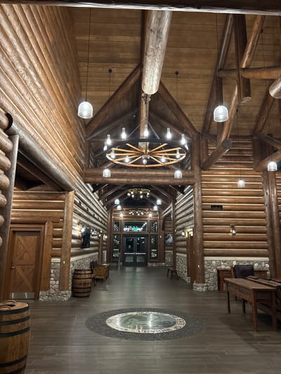 The image depicts the interior of a rustic, lodge-style building, likely a hotel lobby or common area. The architecture features extensive use of natural wood, with thick, round logs forming the walls and ceiling structure. Several wooden beams crisscross overhead, supporting a pitched, wood-paneled ceiling.
Dominating the upper portion of the image is a large, wagon wheel-shaped chandelier with numerous light fixtures. Smaller, globe-shaped pendant lights hang at various intervals throughout the space. The walls are constructed of stacked, horizontally laid logs, with stone bases in some areas.
The floor is a dark, tiled material arranged in a plank pattern. In the center of the floor is a circular mosaic inlay featuring what appears to be a seal with text around its perimeter, possibly indicating a location or organization.
To the left, a wooden door with a crisscross pattern is visible, and a wooden barrel stands next to it. Further down the hallway on the left, more wooden barrels and furniture are present. On the right side of the hallway, several rustic wooden tables and chairs are arranged. In the distance, a set of glass double doors leads to another area, which is dimly lit and appears to have more interior lighting visible.
The overall atmosphere is warm and inviting, with a strong emphasis on natural materials and a traditional, lodge-like aesthetic.
This scene is located at the Old Dougherty House in New Harmony, Indiana, United States.