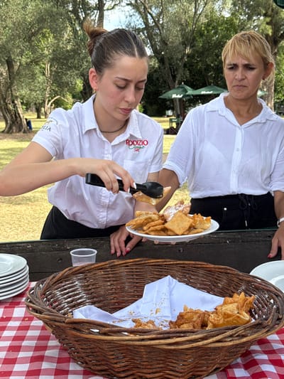 Two women wearing white button-down shirts stand near a picnic table. The younger woman, with a Rodizio Grill logo on her shirt, uses tongs to move fried pastries from a plate. A wicker basket lined with a white cloth is filled with similar pastries. Plates and a disposable cup sit on the checkered tablecloth. Outdoor trees and grass are visible in the background.