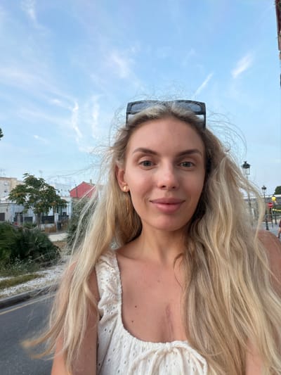 A light-skinned woman with long blonde hair and sunglasses perched on her head smiles at the camera. She is wearing a white eyelet top with ruffled sleeves and a tie in the front. A scar is visible on her chest. The background shows a street with trees and buildings, and a stop sign is visible in the distance. The location is the Benimàmet-beniferri district of Valencia, Spain.