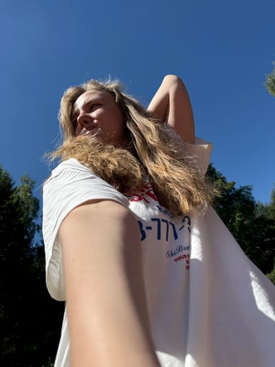A low-angle shot captures a person with long, wavy blonde hair looking upwards and to their left. Their right arm is bent, with the hand positioned behind their head. They are wearing a white short-sleeved t-shirt with partial text visible, including "3-771-3" (likely a phone number) and "Ski Bum," followed by fragments such as "YOUR P," "GI," and "AP." An outstretched arm, possibly the subject's left arm holding the camera, partially obscures the lower-right foreground. The background is dominated by a clear, bright blue sky, with dark green foliage from trees visible along the left and right edges of the frame, indicating an outdoor setting under direct sunlight. The image provides no visual indicators to confirm the specific location within the city of Копнино, Russia.