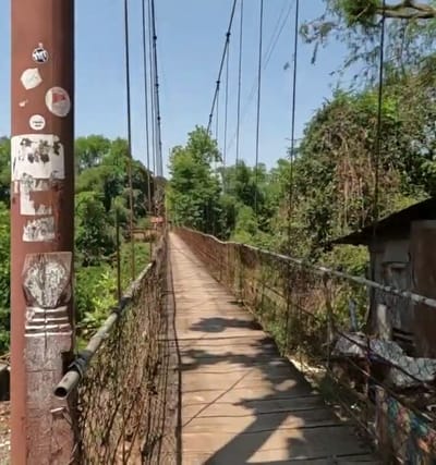 A first-person perspective shot captures a wooden suspension bridge stretching into the distance, flanked by lush green foliage and trees. The bridge's wooden planks are worn, and its metal railings are weathered and rusted. On the left side of the bridge, a thick, reddish-brown metal support post is visible, adorned with several stickers. The sky is a clear blue. On the right side of the bridge, a small, dilapidated structure with a tin roof is partially visible. The scene appears to be set in a rural or natural environment, likely in or around Krong Battambang, Cambodia, given the vegetation and the style of the bridge, which are common in such areas. There are no people or animals visible in the image.