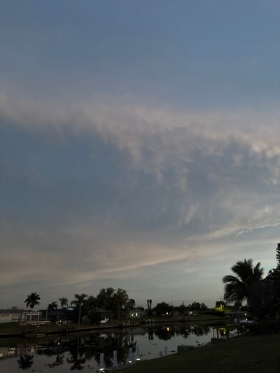 This is a photograph of a canal in North Port, Florida. The sky is a pale blue with wispy, pink-tinged clouds. The canal water is dark and reflects the houses and palm trees that line its banks. The houses appear to be single-family residences with manicured lawns. There are no people visible in the photograph. The overall scene suggests a peaceful evening in a suburban waterfront community.